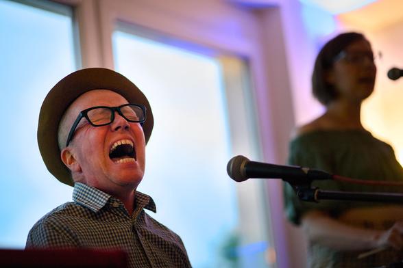 Ein Sänger mit Hut und Brille singt mit geschlossenen Augen, weit auf gerissenem Mund und intensivem Gesichtsausdruck in ein Mikrofon. Im Hintergrund eine weitere Musikerin.