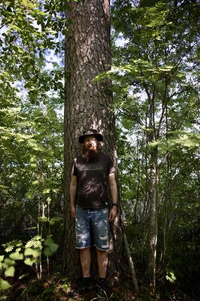 Kilpikaarnainen mänty ja männyn edessä t-paitaan ja risaisiin farkkushortseihin pukeutunut risupartainen ihminen pönöttämässä. Ihmisen hartialeveys ei riitä peittämään puuta. Puun ympärillä pusikkoists rantaa.