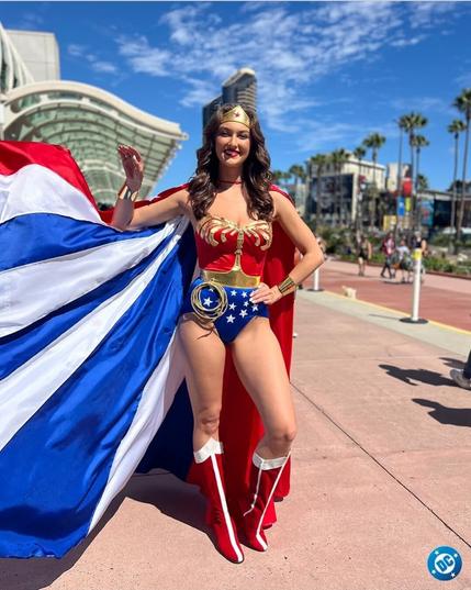 A lovely smiling lady is standing on a sunny street in San Diego. Her name is Rachel Pizzolato. She is wearing a costume identical to that worn by actress Linda Carter in promotional photos for the 1970's Wonder Woman TV series and which was also used in episode 108, "The Last of the Two-Dollar Bills."

The traditional 1970s red and blue Wonder Woman bodice piece is highlighted with white stars on the blue shorts section and gold inlay on the red top half, with a gold belt between them. She has red boots with white trim, a metal tiara, and metal bracelets (presumably made of Feminum). She also has a huge cape in a design reminiscent of the American flag, with long wide vertical stripes of white and blue, with a couple of red stripes among them. Someone off-camera is holding the cape open to show it better. 

Photo copied from the Official DC Comics Facebook account.
