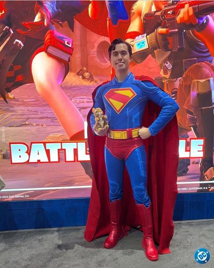 A man is standing in front of a large poster. He is dressed like the newest David Corenswet version of Superman, including the same style of S on his chest. His costume is incredibly accurate and well-done, matching colors perfectly. His physique also matches the character well. He is standing proudly with one hand on his hip, and in the other arm, he is holding a small stuffed squirrel (in keeping with a scene from the 2025 Superman movie, where Superman saves a squirrel in the middle of a battle). 

Photo copied from the Official DC Comics Facebook account.