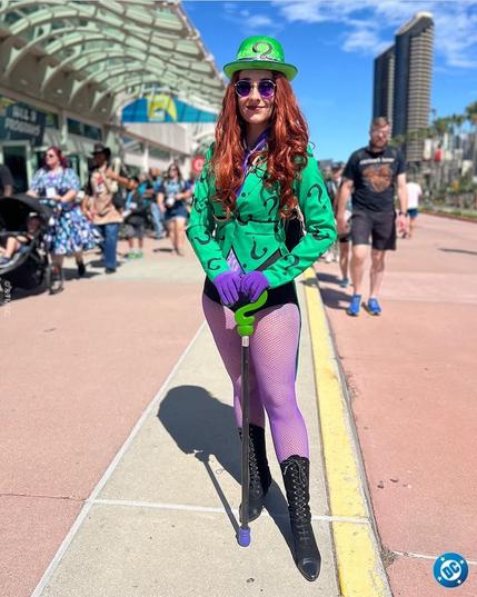 Photo taken on a sunny street in San Diego. A young slim woman with long red hair is dressed like The Riddler. She has tall black boots, purple tights, short black shorts, and a long-sleeve green shirt with question marks on it. She is wearing a rounded green bowler hat and holding a cane with a question mark as the handle. The shades of green and purple used here match the animated Batman colors for the Riddler character very well. 

Photo copied from the Official DC Comics Facebook account.