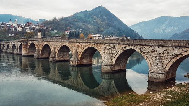 A wide, horizontal shot captures the Mehmed Paša Sokolović Bridge spanning a reflective river under an overcast sky. The medieval stone bridge, characterized by its numerous arches, stretches from the right foreground towards the left background. On the left bank of the river, a town with terracotta roofs and white buildings, is nestled against the base of a verdant hill. In the background, a large, tree-covered mountain rises, its peak just visible against the cloudy sky. The green and blue river reflects the bridge and the cloudy sky, creating a mirrored effect.