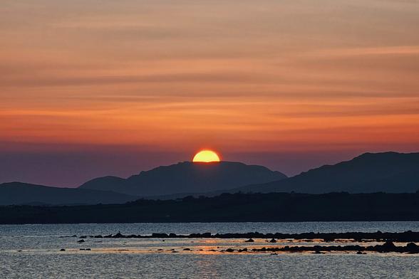 A breathtaking sunset over a body of water, with the sun, a vibrant orange orb, dipping behind a range of dark mountains in the distance. The sky is awash with warm orange, pink, and purple hues, reflecting gently on the calm water where dark rocks are visible near the shore. The overall mood is serene and awe-inspiring.