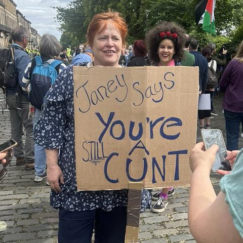 A Scottish woman holds a cardboard sign reading "Janey Says You're STILL A CUNT" at a rally, surrounded by a crowd of demonstrators.