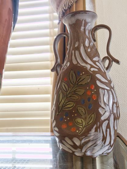 A brown pottery vase with two curving handles stands on a glass surface near window blinds. The vase features hand-painted white swoops and leafy green branches, dotted with orange, blue, and cream accents. Soft daylight filters through the blinds, highlighting the vase’s matte, earthy texture and organic patterns as light casts gentle reflections on the glass below.