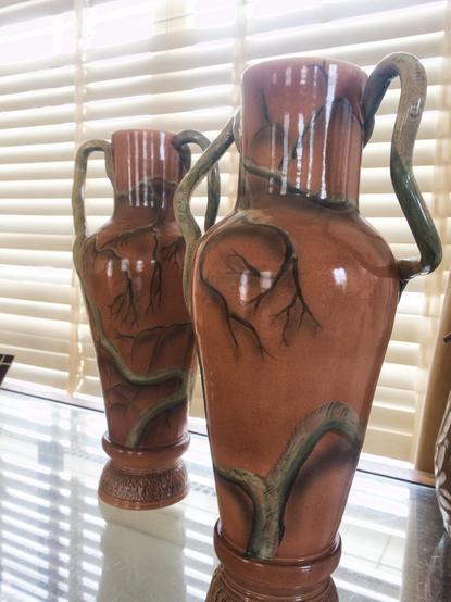 Two tall earthenware vases with double handles stand side by side on a reflective glass shelf, their surfaces glazed in warm terracotta. Each vase is adorned with painted winding green-brown branches, their shapes echoed by the flowing handles. Filtered sunlight from the blinds gives the scene a warm, softly diffused look, accentuating the sheen of the glaze and the vases’ sculptural curves.