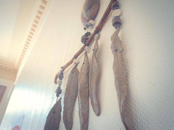 A handmade wall hanging features a series of elongated, sculpted feathers suspended from a curved wooden branch. Each feather, detailed with delicate ridges, hangs from cords alongside interspersed round and disc-shaped beads. The view is angled upward, set against a softly lit, cream-textured wall, with diffused daylight casting gentle highlights and subtle shadows along the wall and ceiling molding.