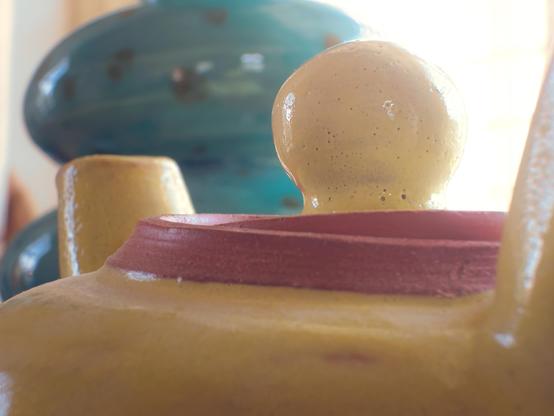 A tight crop focuses on the maroon-lipped opening and round yellow knob atop the yellow teapot. The ceramic surface is dotted and lightly speckled, with the turquoise vase behind lending cool color contrast. Soft highlights and shadows curve across the glossy glaze, bringing out the tactile quality of the piece.