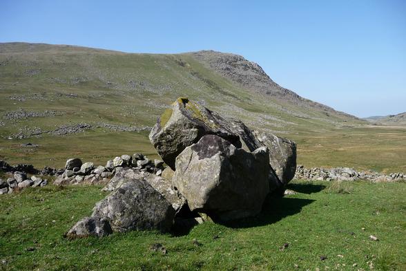 Megaliths potentially forming the remains of a collapsed chamber tomb. A large slab is propped diagonally, supported by a large block. The site is at the head of a valley beneath mountain ridges. A high ridge blocks the views across the centre skyline. The valley drops away gently to the right. It's very sunny and the sky is blue.