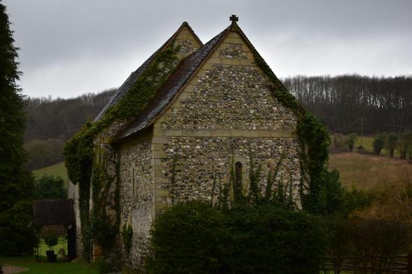 A weathered stone church with flint walls and moss-covered roof sits in a rural countryside setting. The medieval building features distinctive triangular gables and a small bellcote, with ivy and vegetation growing up its walls. Rolling hills and bare winter trees form the backdrop, whilst a graveyard with headstones is visible to the left. The overcast sky and muted lighting suggest a typical English countryside scene on a grey day.​​​​​​​​​​​​​​​​