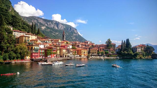 A picturesque panoramic view of the colourful village of Varenna on the shore of Lake Como, Italy. The vibrant terracotta, yellow, and red buildings with green shutters are nestled against a lush green mountainside under a bright blue sky with scattered white clouds. Several boats are dotted across the deep blue lake, with people enjoying the sunny weather. To the left, a lone red kayak adds a splash of contrast. The towering mountains in the background complete this idyllic European scene.