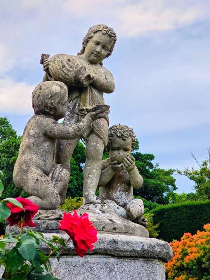 A weathered stone statue group of three cherubic figures stands amidst lush garden foliage under a soft, cloudy sky. In the foreground, vibrant red roses bloom, adding a striking splash of colour to the scene. The central cherub stands holding a large jug, while another kneels, reaching towards it, and a third sits, hands clasped to its face. The statues are covered in moss and lichen, indicating age and adding to their classical charm.