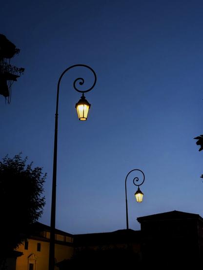 En una vista del cielo nocturno, destacan dos farolas altas. Los postes que sujetan cada farol tienen un diseño delicado, como de un zarzillo de una planta que se enrosca sobre sí mismo en una espiral.

Los faroles destacan con su luz amarilla en el cielo de un azul oscuro.

La imagen transmite mucha calma.