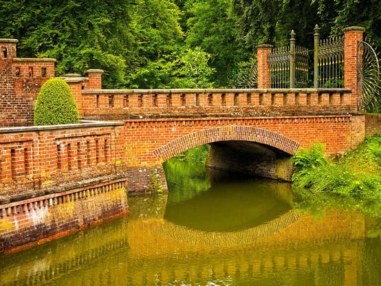 How do I describe this picture so that it becomes clear why it is a perfect Silent Sunday photo for me? There are lots of trees in the background of the photo. All wearing their green summer dress. In front of them is a straight bridge made of red brick. It's very symmetrical, which I like. You can see a gate made of bars on the right. The centre part of the gate is open. There are brick pillars to the left and right of the gate. The gate looks delicate. If you cross the bridge, you come across even more brick. There is water under the bridge. It is green with algae. The bridge is reflected in it. A very peaceful, calm, warm picture.