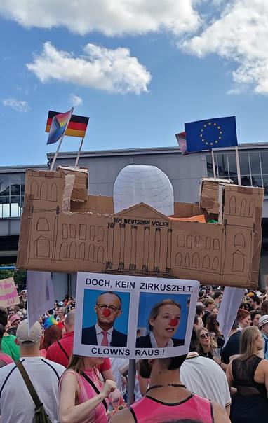 A protest sculpture at CSD Berlin, held up against the backdrop of the above street level Nollendorfplatz metro station. It's a carton replica of the Bundestag building (German Federal Parliament) with flags on its four towers. A printout is taped to the bottom of it showing the faces of German chancellor Friedrich Merz and leader of the far-right/neo-fascist AfD, Alice Weidel, each with a red clowns nose superimposed. It reads "Doch, Kein Zirkuszelt" alluding to a quote of Merz, turning its original negation into a reaffirmative negation. His parties president of the Bundestag Julia Klöckner had decreed not to put up a pride flag on the building for the Christopher Street Day in Berlin, which Merz commented with the insinuation that the colorful flag would make it look like the big top of a circus, which he claimed it was not. The rephrasing reaffirms it isn't one, adding at the bottom "Clowns raus!" [Clowns out!], as the ones who are turning it into a circus are right-wing populists, fascists and those who, like Merz, are not trusted to get in liege with fascists like Weidel's AfD he is trying to imitate.

The flags on top of the building are one German, one EU and 2 Progress Pride flags.