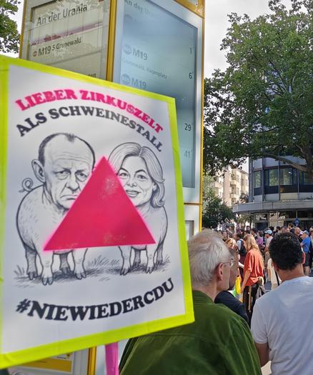CSD Berlin at the bus stop "An der Urania". A protest sign with neon yellow outline and a printed poster of two pigs with the heads of German Chancellor Friedrich Merz and president of the Bundestag Julia Klöckner. For the full context read the previous picture's alt text.

Further than that, Klöckner had also forbidden the rainbow alliance of parliamentarians in the Bundestag to participate in the CSD as representatives of the Bundestag. Both Merz and Klöckner are part of the CDU.

The caption reads: "Lieber Zirkuszelt als Schweine Stall #NieWiederCDU" [Better a big top than a pig sty #NeverAgainCDU], the first part of the sentence is in neon pink, the same color of an upward pointing triangle slapped over the drawing of the two pigs. The pink triangle was the sign stitched onto the clothes of homosexual men in Nazi concentration camps. It has since been reclaimed by gay movements.

Fun fact: A variant with overlapping pink and blue triangles which form a violet triangle in their intersection is used as a sign for bisexuality.