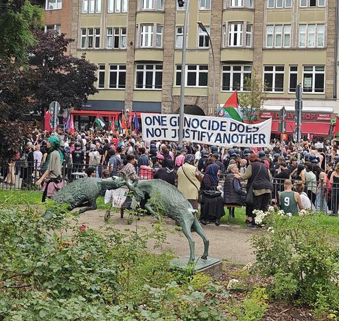 A photo of the Intersectionalist Queer Pride, or IQP for short, as it moves past a park in Neukölln, Berlin. The march is accompanied by a long row of police in riot gear, balaclavas and helmets donned. The police had started slowing the protest down to a crawl just one block earlier, pulling out several individuals before donning their helmets as the vibe shifted towards anger. 

The protestors have a huge banner reading "ONE GENOCIDE DOES NOT JUSTIFY ANOTHER". A Palestinian flag behind it and a few more ahead of it in the parade.

In the foreground the park features a bronce statue of two goats clashing their horns together.

Berlin's police force has been super aggressive against pro Palestinian protests and politicians and newspapers were quick to decry them as "Imported Antisemitism", no matter who partook, but mostly judging it based on the skin color of the participants. To this day, many remain oblivious to how that showcases more of their own blatant racism than the anti-occupation, anti-Isrealian-government and anti-Netanyahu sentiments of the protests.

The IQP decried occupations, displacements and genocides, forced famines and food shortages in Palestine, Kashmir, Congo, Sudan and others. It also protested against rising state violence against trans people worldwide.

Its key statement being: "There is no queer liberation without anti-imperialist, anti-colonial, and anti-Zionist struggle. [...] None of us are free until all of us are free!"
