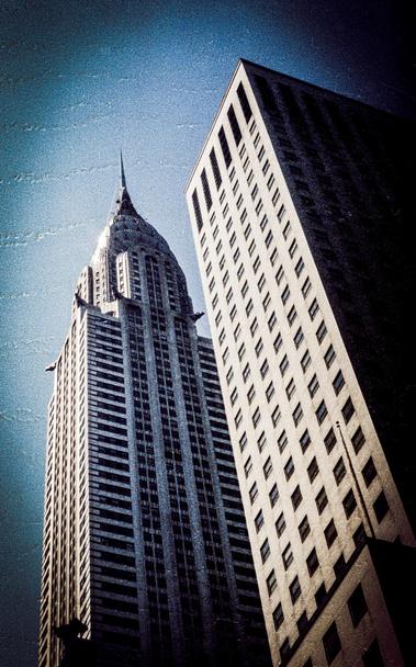 Two skyscrapers rising against a clear blue sky, with the left building featuring an ornate spire and art deco design, while the right building has a simple rectangular shape with multiple windows. The perspective is upwards, capturing both structures from below.