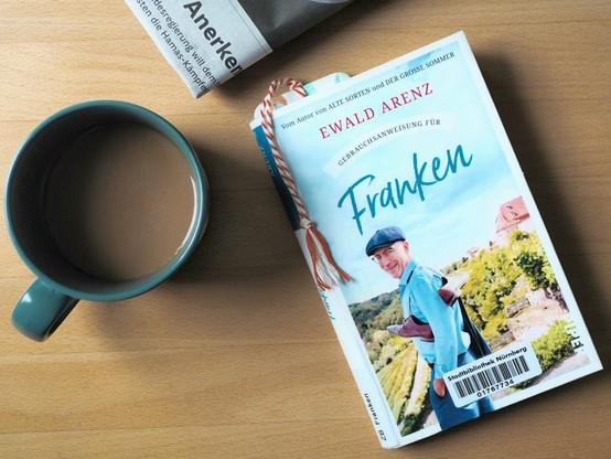 Das Buch "Gebrauchsanweisung für Franken" von Ewald Arenz liegt auf einem Holztisch. Daneben Kaffee in einer dunkelgrünen Tasse. Oben ins Bild ragt die Ecke einer Zeitung.