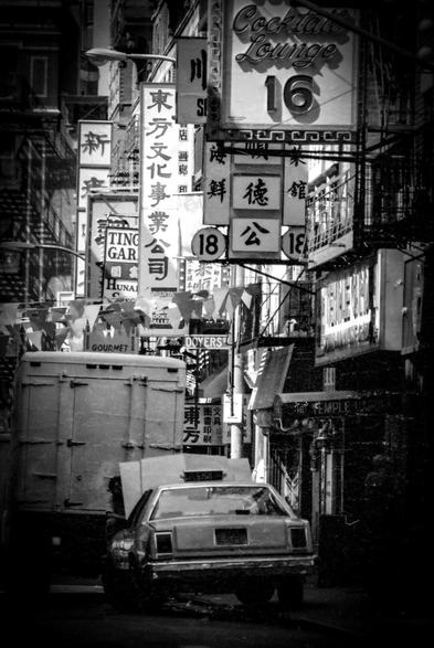 Crowded urban street scene in black and white featuring numerous vertical and horizontal signs with Chinese and English text. A vintage car and a large truck are visible in the foreground, partially obscured by the dense signage above. Overhead, strings of triangular flags hang between buildings. The signs advertise various businesses, including a cocktail lounge. Fire escapes are seen on building facades.