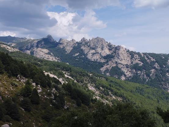 Picos de la Pedriza. Montaña de varios picos rocosos con laderas arboladas. Cielo azul y nubes blancas esponjosas