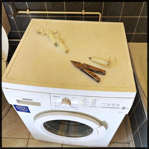 A washing machine in a bathroom. On top are 3 transport security bolts with long screws inside. There is also a Leatherman Multitool unfolded to use the pliers.