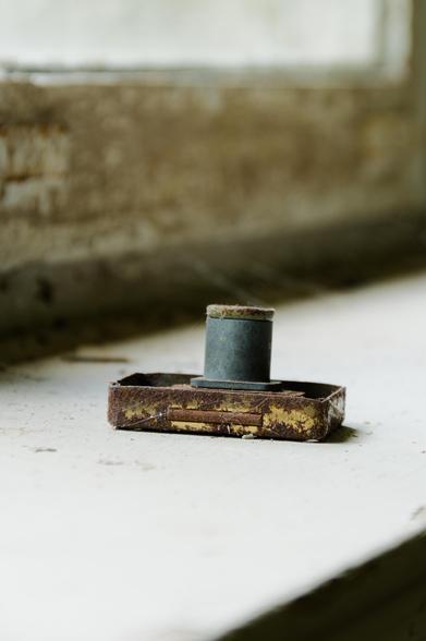 A rusty, old furniture lock lies on a dusty windowsill. In the background, a blurry view of a stained, peeling window frame can be seen. 
###
Eine rostiges, altes Möbelschloss liegt  auf einem staubigen Fensterbrett. Im Hintergrund ist ein verschwommener Blick auf einen fleckigen, abblätternden Fensterrahmen zu sehen.