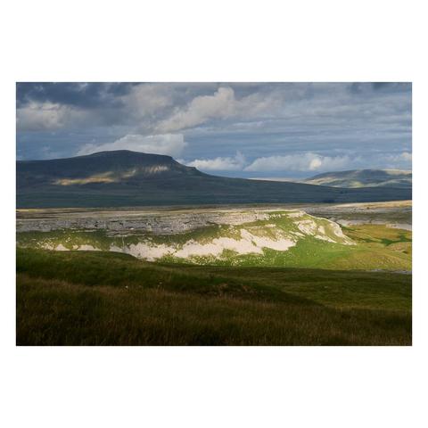 A dramatic landscape view of Crummackdale in the Yorkshire Dales, looking towards the distinctive flat-topped hill of Pen-y-Ghent in the distance. The scene is bathed in soft, moody light as sunlight breaks through heavy, textured clouds, casting patches of brightness on the grassy slopes and limestone cliffs below. The foreground is filled with rich green grass and rolling hills, while the middle ground reveals the pale, weathered limestone escarpments that contrast sharply with the darker, shadowed slopes of Pen-y-Ghent behind. The overall atmosphere is one of rugged natural beauty and changing weather, typical of this iconic English upland area.