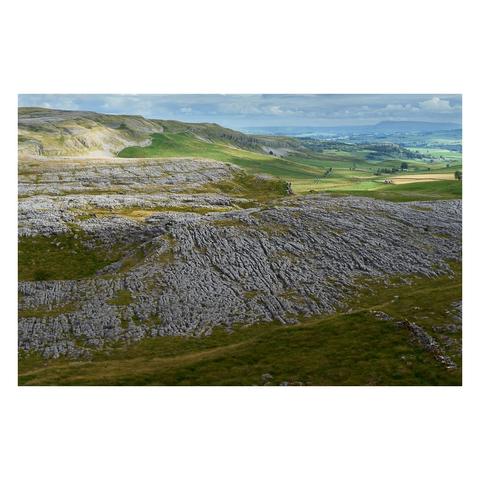 An expansive view from the top of Crummackdale, showcasing the distinctive limestone pavement that spreads broadly across the landscape. The foreground features weathered grey limestone slabs with deep fissures and clints typical of this geological formation, interspersed with patches of grass. Beyond the pavement, the land rises gently to grassy hills and ridges, bathed in dappled sunlight under a partly cloudy sky. In the distance, lush green fields and rolling hills stretch towards the horizon, creating a striking contrast with the rugged limestone. The overall scene conveys the unique natural beauty and geological character of the Yorkshire Dales uplands.
