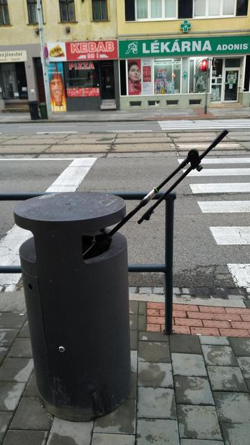 A dark gray trash can next to a crosswalk. Two car lanes and two tram lanes are visible in the background. On the other side of the street, there is a kebab and pizza shop and a pharmacy.