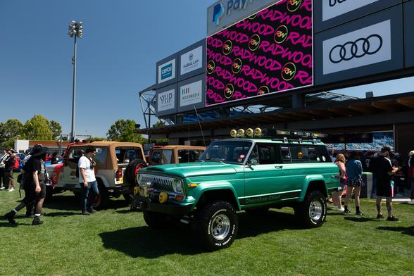 A green vintage lifted SUV and other cars on a grass pitch among spectators, next to a stadium concession area under giant billboards with advertisements. The center billboard display shows purple RADWOOD logos repeated across a black background.