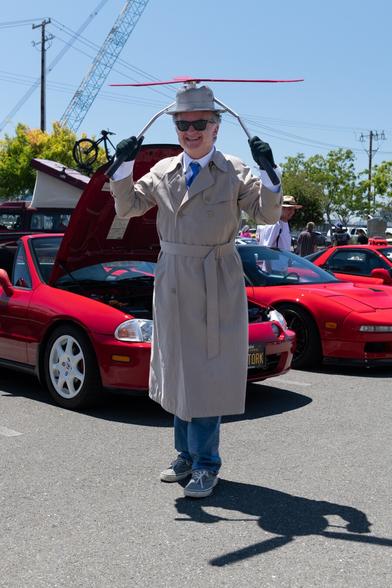 A man in a gray trenchcoat. He wears a matching gray fedora that has a giant propeller mounted on top, and handlebars coming off the sides. His gloved hands are holding the handlebars. He is smiling and posing in front of a row of cars under a blue sky.