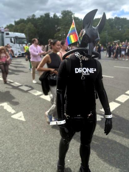 The Jackal Drone on a filled road. Beyond its typical gear, it has a short leash attached to its collar and a rainbow pride flag on its mask. In the background, you can see many different people participating and partying at the gay parade, as well as one of the many stage trucks.