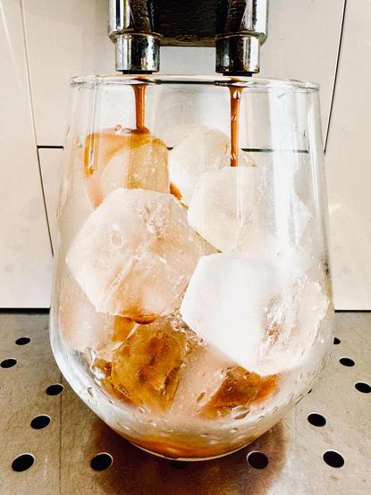 Auf dem Blech einer Kaffeemaschine steht ein Glas voller großer Eiswürfel. Aus der Maschine fließt langsam dunkler, starker Espresso über das Eis.