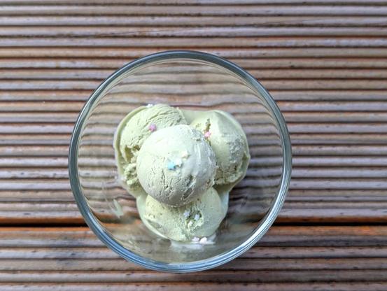 4 Kugeln selbstgemachtes Matchaeis in einer Glasschüssel, fotografiert von oben.
Kleine bunte Zuckersterne als Deko. 
Das Glas steht auf geriffeltem Holz.