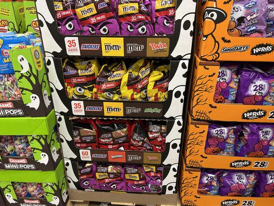 A display of assorted candy bags featuring brands like M&M's, Snickers, Twix, Milky Way, Skittles, SweetTarts, and Nerds. The packaging includes Halloween-themed designs.