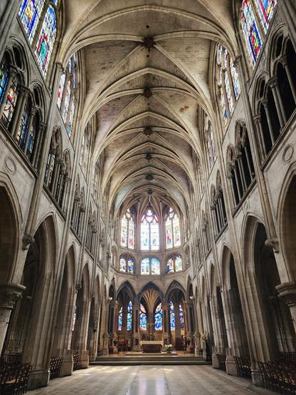 view directly down the central aisle of a gothic cathedral, floor cleared of chairs. The altar with a cross above it is at the focal point, and the glowing stained glass windows and the vaulted arches of the high ceiling take up much of the scene.