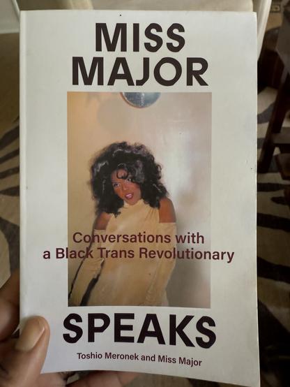 My hand holding a book cover titled "MISS MAJOR SPEAKS: Conversations with a Black Trans Revolutionary" features a photograph of a Black transgender woman. The cover includes the authors' names, Toshio Meronek and Miss Major. The background of the cover is white, the title is in large all-caps San serif font above and below a rectangular photo of Miss Major in her younger years in a shoulderless gown, full make up and long bouncy hair full of waves. The subtitle: “A Black Trans Revolutionary” runs through the middle of the cover.