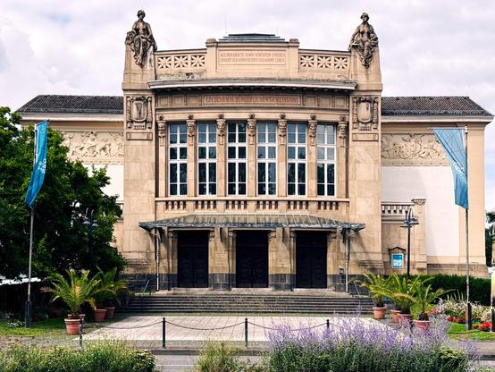 Frontalansicht des Stadttheaters Gießen bei Tageslicht. Das neoklassizistische Gebäude ist symmetrisch aufgebaut, flankiert von zwei Statuen auf den Dachvorsprüngen. Im Vordergrund blüht Lavendel, flankiert von Palmen und Blumenbeeten. Zwei hellblaue Fahnen rahmen die Szene ein.