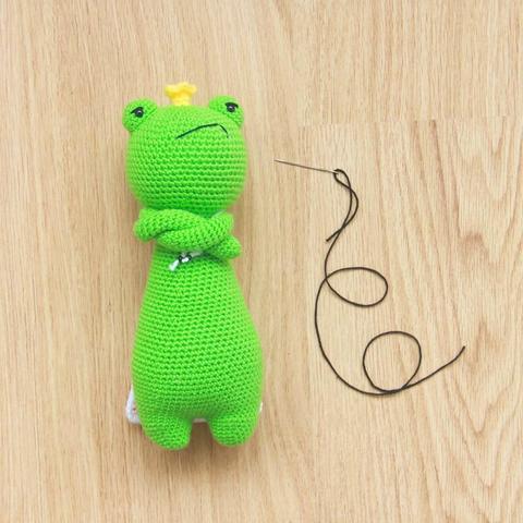 Picture of a crocheted frog with a crown. It's on the floor, arms folded in front of its body and with a very grumpy face. Next to the frog is a sewing needle and some black thread.