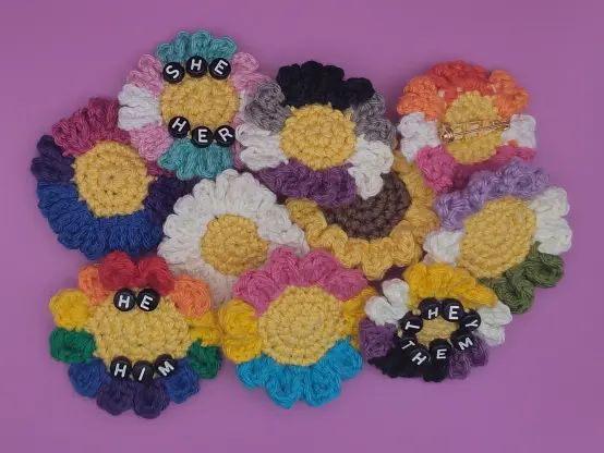 Crochet flower pins stacked on top of each other. In the middle are a daisy and a sunflower, and around them are variant with petals of colors matching various LGBTQIA+ pride flags, rainbow, bisexual, transgender, pansexual, non-binary, asexual, lesbian, and genderqueer. Three of the pins have black beads with white letters, spelling pairs of pronouns: sher/her, he/him, they/them. One pin is turned around, showing the brooch pin on its back.