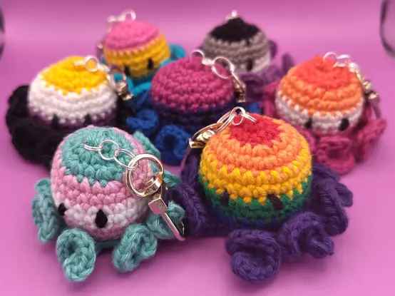 Crochet keychains shaped like octopuses, with colorful stripes matching various LGBTQIA+ pride flags, rainbow, bisexual, transgender, pansexual, non-binary, asexual, and lesbian.