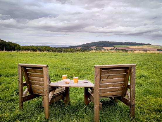 EN: A twin bench stands in the foreground with its back to the viewer, so that the hills and fields that the person sitting there can see are also visible in the picture. On the board between the benches are coffee and brownies still in their packaging, which are sold a few metres away from here.

DE: Eine sog. Twin bench (zwei Holzstühle, die mit einem Holzbrett zu einer Sitzeinheit verbunden sind) steht im Vordergrund mit dem Rücken zum Betrachter, sodass auf dem Bild auch die Hügel und Felder zu sehen sind, die auch der dort Sitzende hat. Auf dem Brett zwischen dem Bänken steht Kaffee und noch eingepackte Brownies, die wenige Meter von hier verkauft werden.