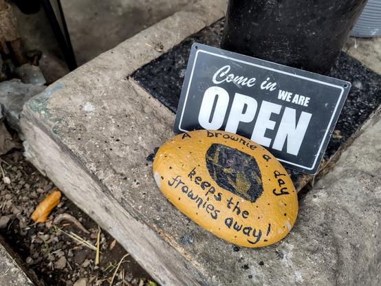 EN: Close-up of the entrance area of the sales barn. A yellow-painted stone bears the inscription ‘A brownie a day keeps the frownies away.’ Behind it is a sign indicating that the shop is open.

DE: Detailaufnahme vom Eingangsbereich der Verkaufsscheune. Auf einem gelb angemalten Stein steht "A brownie a day keeps the frownies away." (Könnte man mit "Ein Brownie am Morgen vertreibt Kummer und Sorgen" sehr frei übersetzen). Dahinter befindet sich ein Schild mit dem Hinweis, dass geöffnet sei.
