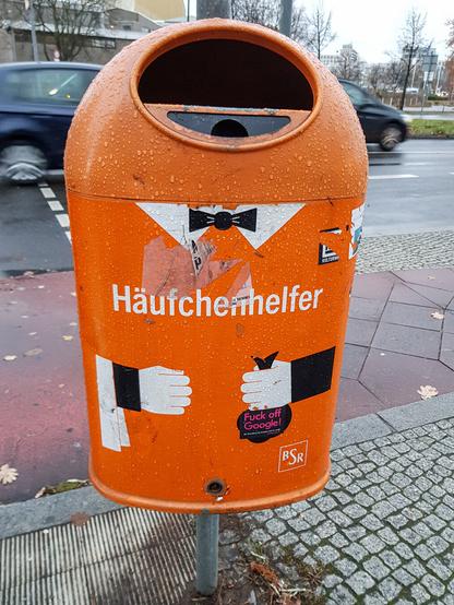 Ein orangener Mülleimer der Berliner Stadtreinigung, der durch drei Aufklebern (z.B. oben ein Kragen mit Fliege) quasi als Butler verkleidet ist. In seiner linken Hand "hält" er einen Hundekotbeutel. Mittig steht das Wort "Häufchenhelfer".
(Außerdem gibt es zusätzliche Sticker. Einer klebt auf dem erwähnten Hundkotbeutel und trägt den Schriftzug "Fuck off Google!"