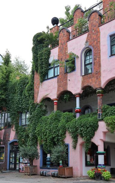 A detailed view of a section of the "Grüne Zitadelle" in Magdeburg, Germany, showcases its vibrant and organic architecture. The building's soft pink facade is heavily draped and interwoven with lush green climbing plants and ivy, giving it a natural, almost overgrown appearance. Curved arches and wavy lines characterise the structure, with sections of reddish-brown brickwork integrated into the pink walls. Distinctive, colourful, and irregularly shaped pillars support various balconies and overhangs, some featuring bright red and blue bands. Numerous windows of differing sizes and shapes are visible, some framed in blue. Planters with small trees and shrubs are placed along the base of the building, which also features dark, arched doorways and ground-floor windows. The sky overhead is a soft grey.