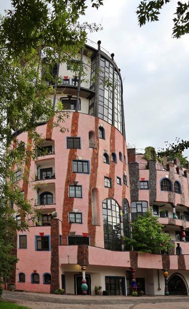 A medium shot of the "Grüne Zitadelle" in Magdeburg, Germany, showcases its whimsical, unconventional architecture. The building's dominant colour is a textured, wavy pink, interspersed with vertical stripes of reddish-brown brickwork. Irregularly shaped windows, some with blue frames, are scattered across the facade. To the right, a prominent, dark-glazed cylindrical tower rises, contrasting with the softer pink tones. Various trees and abundant greenery are integrated into the structure, growing from balconies and the roofline, emphasising its connection to nature. Hints of colourful, playful pillars are visible near the ground floor entrances. The sky is overcast, and green foliage frames the top of the image.