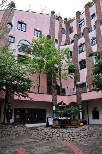 A medium shot captures an inner courtyard of the Grüne Zitadelle in Magdeburg, Germany, revealing its whimsical and organic architecture. The multi-storey building encircles the courtyard, its facade a distinctive wavy pink with integrated vertical bands of reddish-brown brickwork. Irregularly shaped windows, some circular and others arched or rectangular with dark frames, are scattered across the walls. Several trees with vibrant green foliage grow directly within the courtyard, their branches reaching up towards the building, and more greenery is visible on the upper levels.
In the foreground, a round, cobbled plaza features a small, multi-tiered fountain at its centre, surrounded by planters. Colourful, eccentrically shaped pillars, typical of Hundertwasser's style, adorn the building's ground level and support the balconies. A small "INFO" sign is visible above an entrance. The sky is overcast.