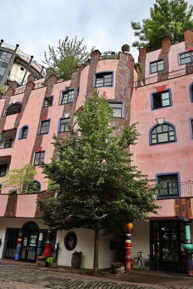 A vibrant, quirky building, the "Grüne Zitadelle" in Magdeburg, Germany, dominates the frame. Its facade is a wavy, organic pink, adorned with irregular windows, some framed in blue. Trees and lush greenery sprout from various levels, including the roof, giving the impression of a natural, living structure. A prominent tree stands in front of the building, its branches reaching towards the wavy pink walls. The architecture is distinctly Hundertwasser-style, characterised by its whimsical shapes and disregard for straight lines. A cobbled path leads to the building's entrance, where colourful, unconventional pillars and a bicycle are visible. The sky above is overcast.