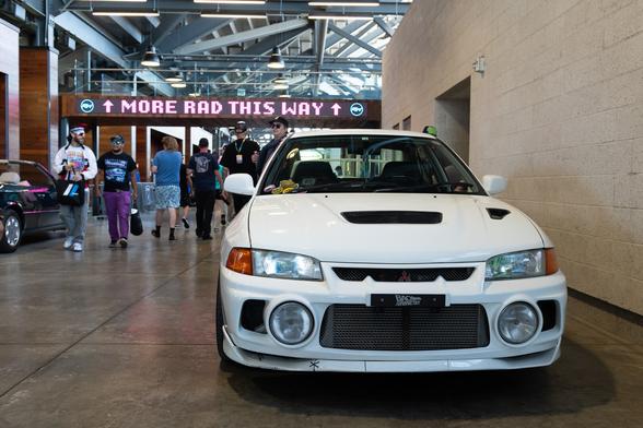 A white Japanese rally sedan in the hallway of a stadium. An LED sign on the gantry reads “MORE RAD THIS WAY”.